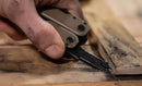 A close-up shows a hand using the Leatherman Super Tool® 300M - Molle Black to pry up a nail from weathered wood, with dust and debris scattered on the surface.