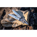 A pair of beige work gloves lies on rocky ground with a Leatherman Surge Multitool on top, partially open to show its pliers.