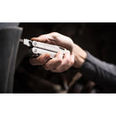 A hand holds a Leatherman Surge Multitool with a screwdriver attachment by a dark surface. The multitool, featuring the signature blade exchanger, stands out in focus against a blurred background.
