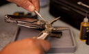 Someone uses a metal tool from the Leatherman Tool Maintenance Kit to clean a multitool’s inner mechanism, with the kit case and a small lubricant bottle nearby on the tray.