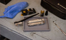 A multitool, cleaning brush, lubricant bottle, blue cloth, cutting tool, and black case from the Leatherman Tool Maintenance Kit are arranged on a gray workbench. The multitool and brush rest on a small tray.