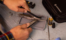 A person cleans a multitool plier with a small brush on a workbench using the Leatherman Tool Maintenance Kit. Nearby are the kit’s Leatherman-labeled case, a bottle of oil, and various small metal tools.