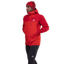 A person in a red Mountain Equipment Mens Lhotse GTX Pro Jacket and matching beanie, with black pants featuring Mountain Equipment logos, stands hands-in-pockets against a white background.