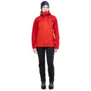A woman modeling a red technical outdoor jacket with a full front zipper, zippered chest pocket, and high collar, paired with black pants and a dark beanie against a plain white background.