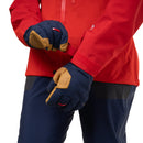 A close-up of a person wearing a red jacket and navy blue pants, adjusting a pair of navy and tan gloves on their hands against a white background.