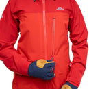 A close-up of a red and orange outdoor jacket with a full front zipper, a vertical chest zip pocket, and a small white Mountaineering brand logo on the left chest. A person's gloved hands are shown holding the zipper area, wearing navy and yellow gloves.