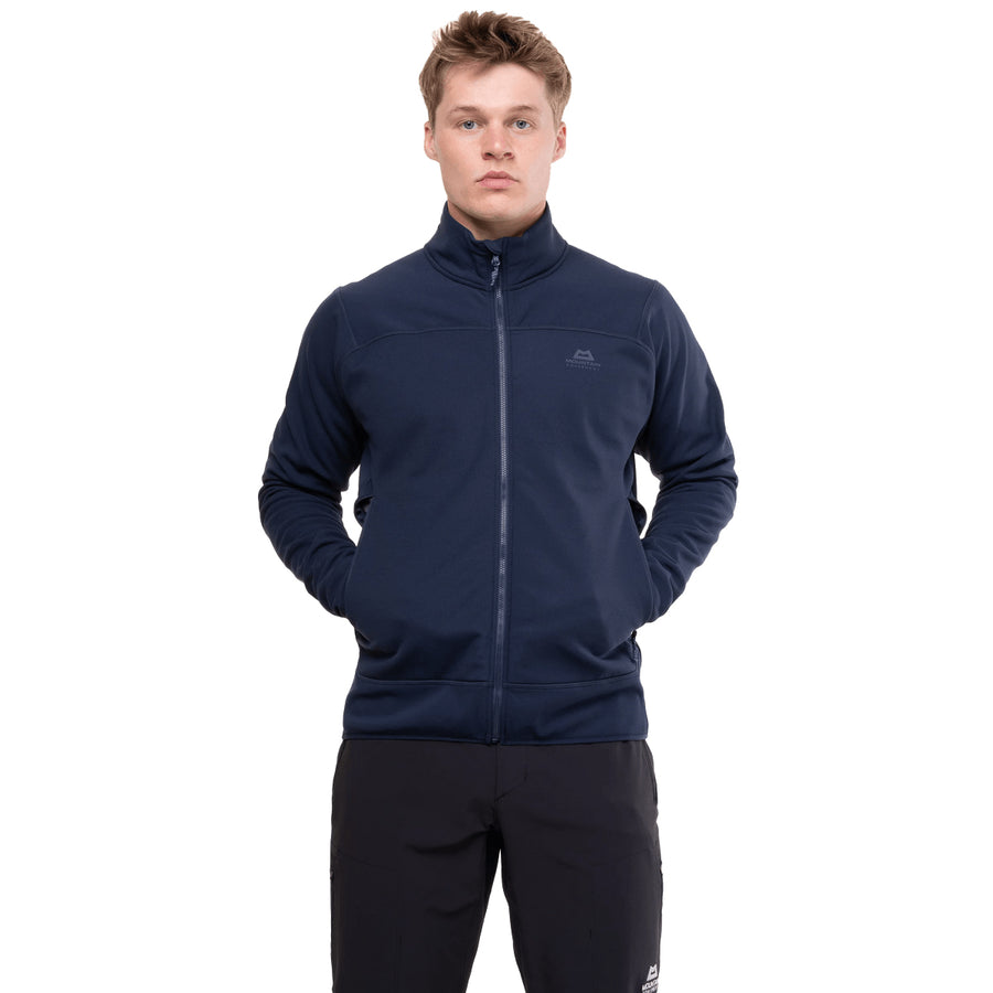 A person stands facing forward, wearing the Mountain Equipment Men’s Jorasses Lite Fleece Jacket in navy blue and black pants, with hands in the jacket pockets against a plain white background.