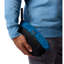 A person wearing a blue sweatshirt and gray pants holds a small, zippered blue and black pouch—with a loop handle—ideal for storing the Columbia Mens Pouring Adventure™ III Waterproof Hiking Jacket.