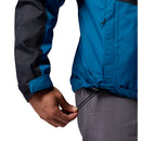 A person wearing a Columbia Mens Pouring Adventure™ III Waterproof Hiking Jacket in blue and black with gray pants adjusts the hem drawstring with their left hand.