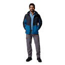 A man stands facing forward with hands in the pockets of his Columbia Mens Pouring Adventure™ III Waterproof Hiking Jacket, worn over a light blue shirt, paired with gray cargo pants and black shoes against a plain white background.