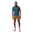 A man stands facing forward, wearing a teal short-sleeve polo shirt, Columbia Mens ROC Tech Shorts in tan, and white sneakers. The background is plain white.