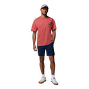 A man stands against a white background wearing a light purple cap, coral pink pocket t-shirt, Columbia Mens ROC Tech Shorts in navy blue, white crew socks, and white sneakers with beige soles. One hand is in his pocket.