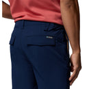 A person is shown from the waist down wearing Columbia Mens ROC Tech Shorts in coral, with a focus on the back pocket featuring a flap and a small Columbia logo tag.