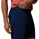 A man wearing Columbia Mens ROC Tech Shorts in dark blue adjusts the waistband with his right hand. The image highlights the shorts’ pockets, stretch comfort, and a small Columbia brand label.