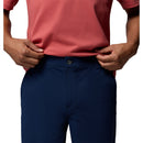 A man models Columbia Mens ROC Tech Shorts in navy blue, holding up a coral short-sleeve shirt to reveal the shorts’ button and waistband against a white background.