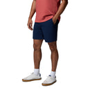 A person stands with hands in pockets, wearing Columbia Mens ROC Tech Shorts in navy blue and a coral t-shirt, paired with white socks and gum-soled sneakers against a plain white background. Their head is not visible in the image.