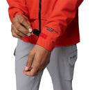 A person adjusts the Velcro wrist strap on the sleeve of a red Columbia Mens Saudan Pro 3L Waterproof Shell Jacket, paired with light gray Columbia pants featuring visible cargo pockets.