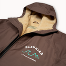 A close-up of a brown hooded jacket with a beige sherpa lining and a front zipper. The left chest features a white and teal graphic logo that reads 'BLEUBIRD' with the tagline 'BE ONE WITH THE ELEMENTS'.