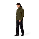 A man wearing a green Rab Mens Namche GTX Waterproof Jacket, black pants, and black shoes stands against a plain white background, looking slightly downward.