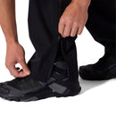 A person zips up the cuff of Rab Mens Namche Hike GORE-TEX Pants over black hiking shoes. Only their hands, lower legs, and shoes are visible against a white background.