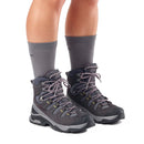 A person wearing Salomon Womens Quest 5 GORE-TEX Hiking Boots with pink laces and mid-calf gray socks stands on a white background, showing only their lower legs and feet in the stylish, waterproof hiking boots.