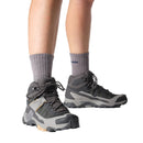 A person stands on a white background wearing Salomon Womens X-Ultra 5 Mid GORE-TEX Hiking Boots in gray and black with a Matryx upper and thick gray socks. The Salomon brand is visible on one sock.