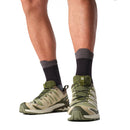A person wearing black socks and Salomon Mens XA Pro 3D V9 GORE-TEX Trail Shoes in green with beige accents, designed for technical terrain, stands on a white background. Only their lower legs and feet are visible.