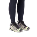 A person wearing dark leggings and Salomon Womens XA Pro 3D V9 GORE-TEX Trail Shoes—black, grey, and light green, designed for technical trails—stands against a white background. Only their lower legs and feet are visible.