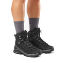 A person wearing gray crew socks and black Salomon Men’s Quest 5 GORE-TEX Hiking Boots stands on a white background, showing only their legs from the knees down.