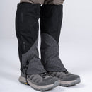 A person wearing gray hiking shoes and black Trekmates Cuillin GORE-TEX Gaiters over light brown pants, standing on a white background. The waterproof Trekmates gaiters cover the lower legs and tops of the shoes, providing outdoor protection ideal for mountain walking.