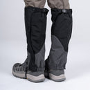 A person stands on a plain light background, wearing gray hiking shoes, gray pants, and Trekmates Cuillin GORE-TEX Gaiters in black and gray, which cover the lower legs and tops of the shoes—perfect for mountain walking.