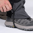 A person fastens a black strap on their grey Trekmates Cuillin GORE-TEX Gaiter over grey hiking boots with a rugged sole, preparing for a day of mountain walking.