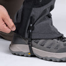 Outfitted in Trekmates Cuillin GORE-TEX Gaiters, a person adjusts the secure closing flap over a hiking boot featuring a gray sole and black-and-gray uppers. The black and gray waterproof gaiter is fastened with its strap under the boot—ideal protection for mountain walking in wet conditions.