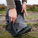 A person outdoors pulls a Trekmates Cuillin GORE-TEX Gaiter over their hiking boot and pant leg, preparing for mountain walking on a rocky, grassy path.