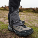 A close-up of a person’s lower leg wearing Trekmates Cuillin GORE-TEX Gaiters and gray hiking boots, standing on a grassy, rocky trail outdoors—ideal for mountain walking.
