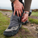 A hiker secures their Trekmates Cuillin GORE-TEX Gaiter over a boot on a rocky trail, clad in a fleece jacket and wristwatch. Grass and stones line the path, emphasizing the essential gear for comfortable mountain walking.