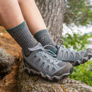 Close-up of a person’s feet in gray hiking shoes and Darn Tough Womens Hiker Micro Crew Midweight Hiking Socks, resting on a tree trunk in the forest. These socks offer a blister-free fit for any outdoor adventure.