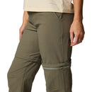 A woman wears Columbia Womens Leslie Falls Hiking Convertible Trousers, showing olive-green zip-off pants with Omni-Shield tech and a beige top; the partially unzipped legs reveal how the pants easily convert into shorts.
