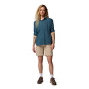 A person with long, wavy blond hair wears a blue Columbia shirt, Columbia Womens Leslie Falls Hiking Convertible Trousers, white socks, and brown shoes, standing and facing forward against a plain white background.