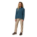 A woman with long blonde hair faces forward in a blue Columbia shirt, Columbia Womens Leslie Falls Hiking Convertible Trousers with Omni-Shield, and hiking shoes, against a plain white background.