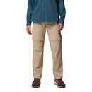 A person wears Columbia Women’s Leslie Falls Hiking Convertible Trousers, featuring zip-off legs to convert into shorts, paired with a teal long-sleeve shirt and hiking shoes. The focus is on the versatile beige pants.
