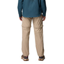 A person stands facing away, wearing Columbia Womens Leslie Falls Hiking Convertible Trousers and a teal long-sleeve shirt with brown hiking shoes. These outdoor-ready trousers feature Omni-Shield technology for durability and comfort.