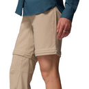A person wears Columbia Womens Leslie Falls Hiking Convertible Trousers—one leg zipped off as shorts, the other full-length—paired with a teal long-sleeve shirt. A gold ring is visible on their hand.