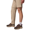 A woman stands against a white background wearing Columbia Womens Leslie Falls Hiking Convertible Trousers with one pant leg zipped off, paired with hiking shoes and white socks, highlighting their versatility and Omni-Shield technology.