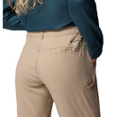 A woman wears a dark blue long-sleeve shirt and light beige Columbia Womens Leslie Falls Hiking Convertible Trousers with Omni-Shield tech, hand in pocket. The image highlights the back and side of the pants and shirt.
