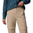 A person wearing a teal long-sleeve button-up shirt and Columbia Womens Leslie Falls Hiking Convertible Trousers stands with one hand on their waist. The beige pants feature zip-off legs and Omni-Shield technology for protection.
