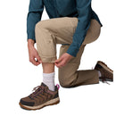 A person kneels, rolling up the cuff of Columbia Womens Leslie Falls Hiking Convertible Trousers, wearing a teal long-sleeve shirt, white socks, and brown hiking shoes.