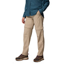 Wearing Columbia Womens Leslie Falls Hiking Convertible Trousers in beige, which zip off at the knee, a teal long-sleeve shirt, and brown hiking shoes, this person stands hands-in-pockets; head not visible.