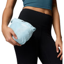 A person in black leggings and a teal crop top holds a compact, folded Columbia Womens Pouring Adventure III Waterproof Hiking Jacket in light blue against their hip.