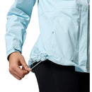 A person adjusts the hem drawcord of a light blue Columbia Womens Pouring Adventure III Waterproof Hiking Jacket, paired with black pants. Only the torso and hand are visible.
